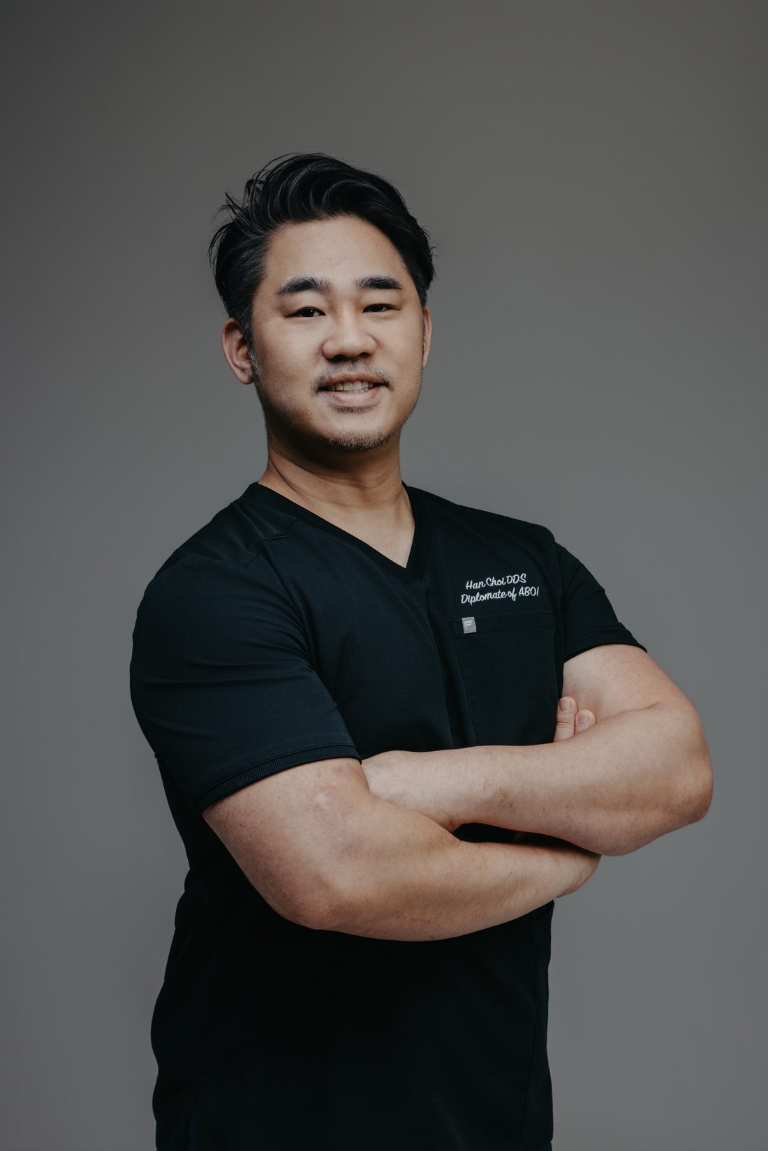Dr. Han Choi in black scrubs embroidered with his name and ABOI diplomate title, arms crossed, against a grey studio backdrop.